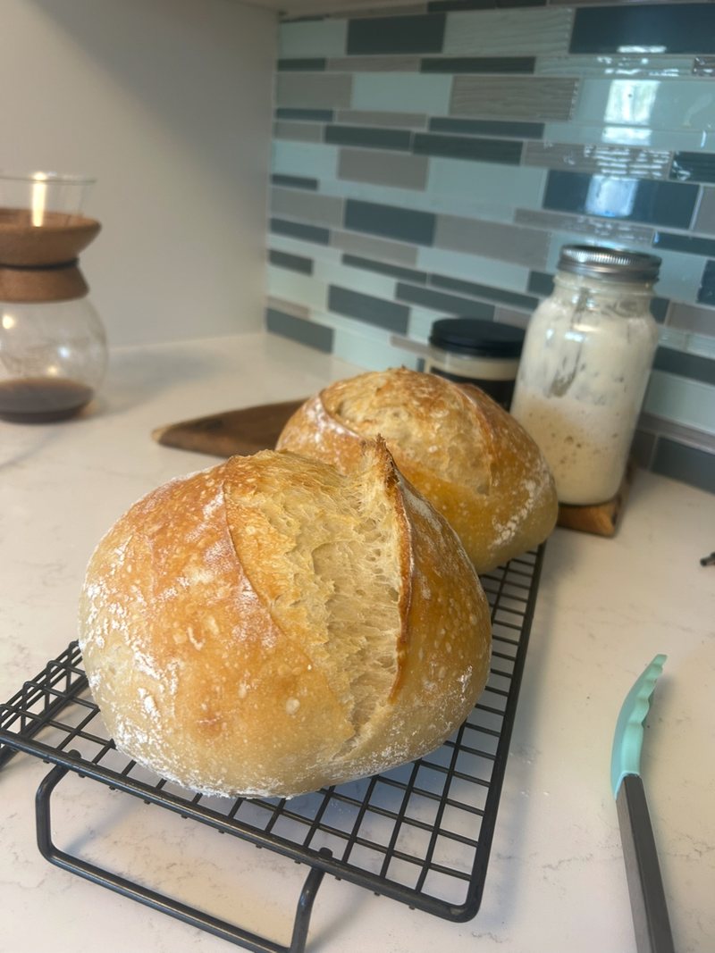 Two round sourdough loaves cooling on a wire rack, with a jar of starter behind them
